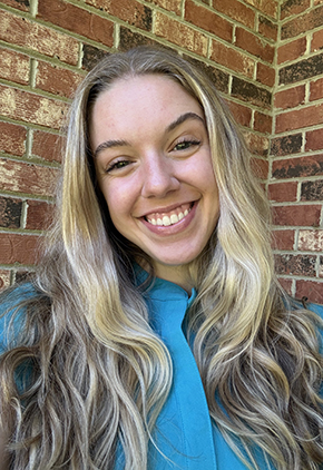 Dr. Hallie Braeuner smiles outside in front of a brick building. She is wearing blue scrubs.
