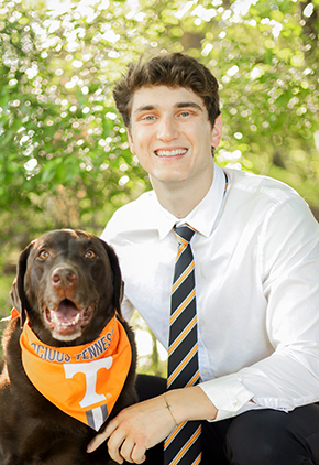 Dr. Brandon Burgess smiles outside while kneeling next to his chocolate Labrador. He is wearing a white dress shirt, black dress pants and a University of Tennessee tie.