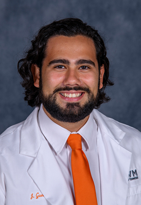 Dr. Jesus Guevara smiles for a professional headshot in front of a grey-blue background. He is wearing his University of Tennessee whitecoat, a white dress shirt and Tennessee orange tie.