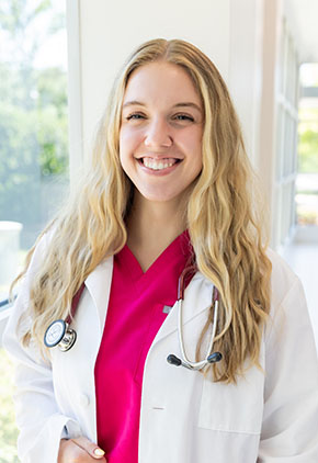 Dr. Hallie Braeuner smiles in the windowed lobby of BluePearl Pet Hospital. She is wearing hot pink scrubs, her whitecoat and has a stethoscope around her neck.
