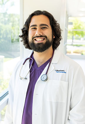 Dr. Jesus Guevara smiles in the windowed lobby of BluePearl Pet Hospital. He is wearing purple scrubs, his whitecoat and has a stethoscope around his neck.