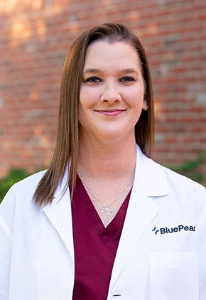 Dr. Nikki Bernard smiles outside in front of a brick building. She is wearing maroon scrubs and her BluePearl whitecoat.