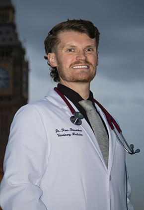 Dr. Ross Brusenhan smiles outside on a cloudy day in London. He's wearing his whitecoat and has a maroon stethoscope around his neck. Big Ben is featured in the background.