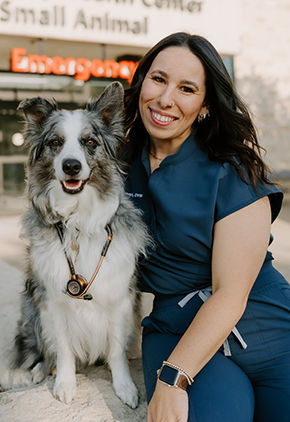 Dr. Meredith Edelmon smiles outside of a veterinary emergency hospital with her Australian shepherd. She is wearing blue scrubs and her dog has a stethoscope around their neck.