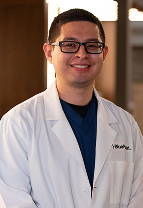 Dr. Daniel Gomez smiles for a headshot in the lobby of BluePearl Pet Hospital. He is wearing a navy shirt and his whitecoat.