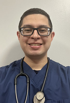 Dr. Daniel Gomez smiles for a headshot in front of a tan background. He is wearing navy scrubs and has a stethoscope around his neck.