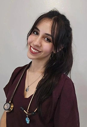 Dr. Beatriz Gutiérrez smiles for a headshot in front of a tan background. She is wearing maroon scrubs and has her stethoscope around her neck.