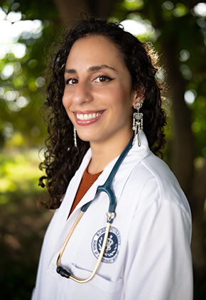Dr. Jessica Karkar smiles outside for a professional headshot. She is wearing a red top and her Ross University whitecoat with a stethoscope around her neck.