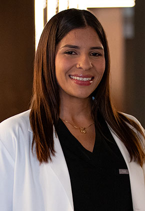 Dr. Kimberley Savinovich smiles for a professional headshot in the lobby of BluePearl. She is wearing black scrubs and her BluePearl whitecoat.