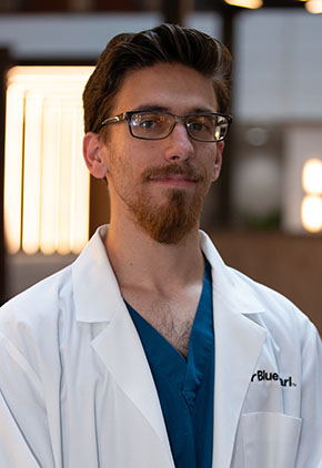 Dr. Stephen Zachary Trombetta has a soft smiles as he poses for a headshot in the lobby of BluePearl Pet Hospital. He is wearing turquoise scrubs and his whitecoat.
