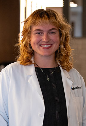Dr. Sophia Janssen-Madden smiles for a headshot in the lobby of BluePearl. She is wearing a black top and her whitecoat.
