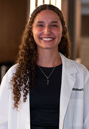 Dr. Zoe Kelly smiles for a headshot in the lobby of BluePearl. She is wearing a black top and her whitecoat.