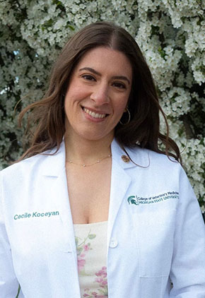 Dr. Cecile Koceyan smiles outside in front of a tree. She is wearing a white floral dress and her Michigan State whitecoat.