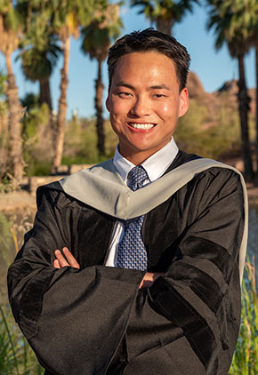 Dr. Christopher Minami smiles outside in front of palm trees. He is wearing his doctoral robe.