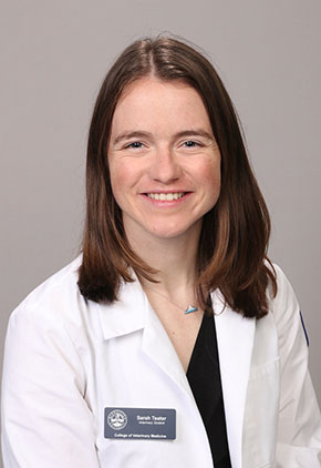 Dr. Sarah Teter smiles for a professional headshot in front of a tan backdrop. She is wearing a black top and whitecoat with her nametag.