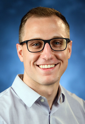 Dr. Seth Locker smiles for a professional studio headshot in front of a blue background. He is wearing a light blue dress shirt and black glasses.