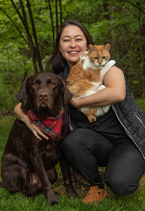 Dr. Rachel Natsume smiles outside while holding her orange cat and kneeling with one arm around her dog. She is wearing black pants, blouse and vest.