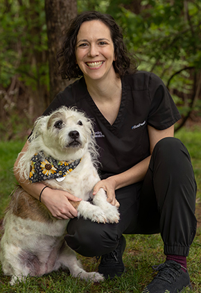 Dr. Natalie Fording smiles outside in a lush wooded area. She is wearing black scrubs and kneeling next to her white dog.