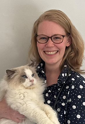 Dr. Zoe Fautz smiles while holding up her white, fluffy cat. She is wearing a navy shirt with white polka dots.