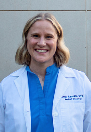 Dr. Emily Lemoine smiles outside in front of a building wall. She is wearing a blue blouse and her whitecoat.