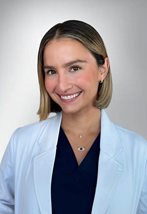 Dr. Valeria Benítez-Vera smiles for a headshot in front of a white backdrop. She is wearing black scrubs and her whitecoat.