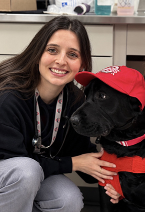 Dr. Stephanie Shemilt kneels next to a black Labrador in the triage area of the hospital. She is wearing a black jacket and grey scrubs.