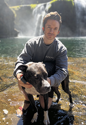Dr. Jacob Franklin smiles outside, kneeling in the pool of a waterfall. He is hugging his brindle pit bull.