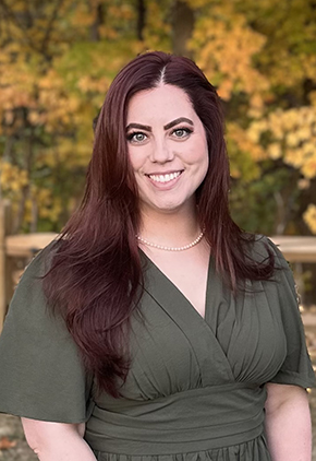 Dr. Michaela Clouden smiles outside in front of fall foliage. She is wearing an olive green blouse.