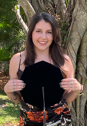 Dr. Emily Rothberger smiles outside in front of a tree. She is wearing a black top, floral skirt and is holding her doctoral cap in front of herself.
