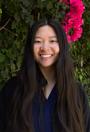 Dr. Cynthia Tom smiles outside in front of a flowering tree. She is wearing black scrubs and a black jacket.