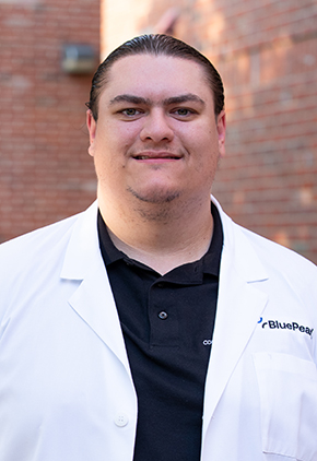 Dr. Matt Elbe smiles outside in front of a brick building. He is wearing a black polo and his BluePearl whitecoat.