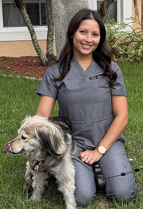 Dr. McKenna Hernandez smiles outside while kneeling next to a young dog. She is wearing grey scrubs.