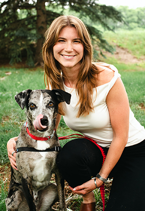 Dr. Kendall Barnes kneels next to her black and white short-haired dog in a open green space. She is wearing black pants and white blouse while her dog has a red leash attached to their harness.