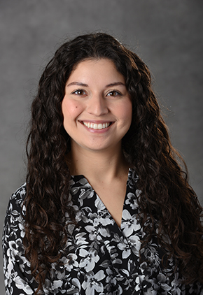 Dr. Victoria Caballero smiles for a professional studio headshot in front of a grey background. She is wearing a black and white blouse.