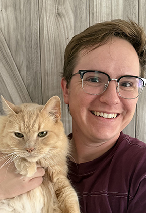 KB Brown smiles in front of a wood wall. They are holding their orange cat and wearing a maroon shirt.