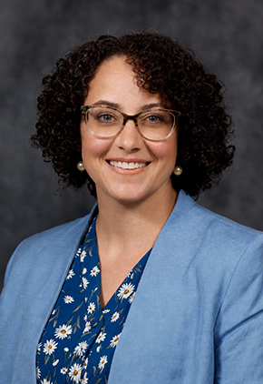 Dr. Janis Lapsley smiles for a professional studio headshot in front of a grey background. She is wearing a blue top that features white daisies and a light blue blazer.