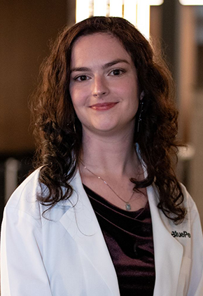 Dr. Sydney Campbell smiles for a professional headshot in the lobby of BluePearl Pet Hospital. She is wearing black scrubs and her whitecoat.