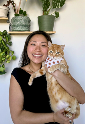 Dr. Caitlin Yung smiles while holding her orange cat up in her arms. They are in front of a white wall that features multiple plants on individual shelves. She is wearing a black blouse.