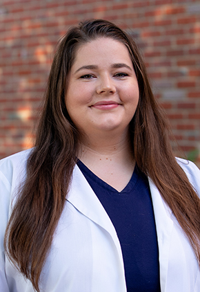 Dr. Megan Walls smiles outside in front of a brick building. She is wearing navy scrubs and her whitecoat.