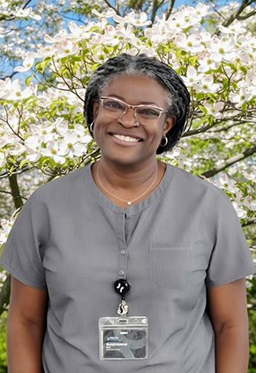 Dr. Bisoye Adedeji smiles outside in front of a tree blooming with little white flowers. She is wearing grey scrubs and has her BluePearl page clipped to the front.