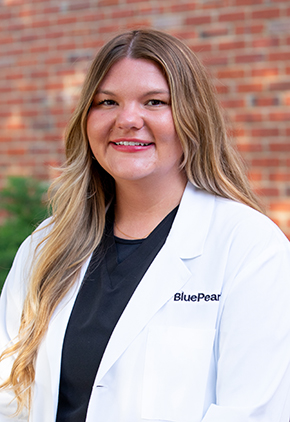 Dr. Koedi Lawley smiles outside in front of a brick building. She is wearing black scrubs and her BluePearl whitecoat.